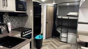 Cocina americana compacta con estufa, fregadero, microondas y horno. Incluye un cubo de basura y una cama litera con almohada.