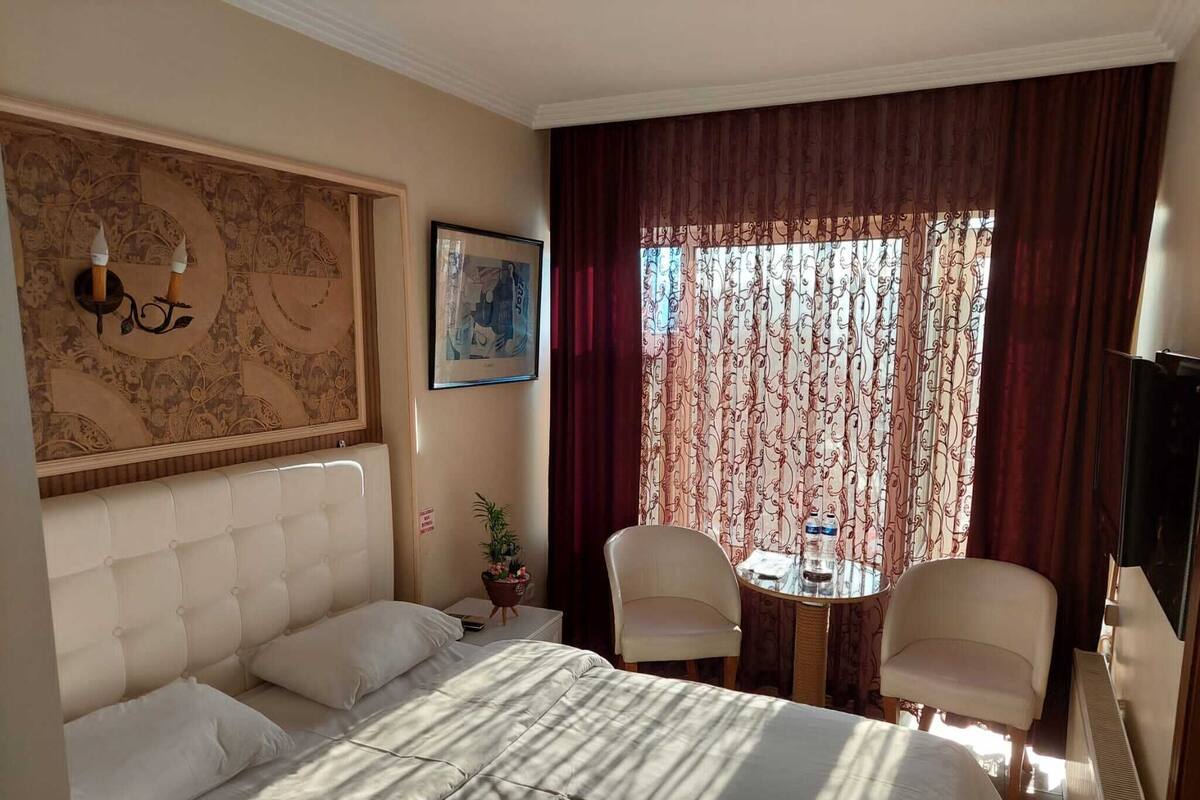 Habitación de hotel con una cama grande, dos sillas, una mesita con botellas de agua, un mapa en la pared y una ventana con cortinas estampadas.