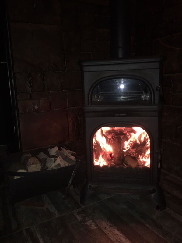 A wood-burning stove with a fire inside, set against a brick wall. - GOLDEN ELİT OTEL (Bandirma)