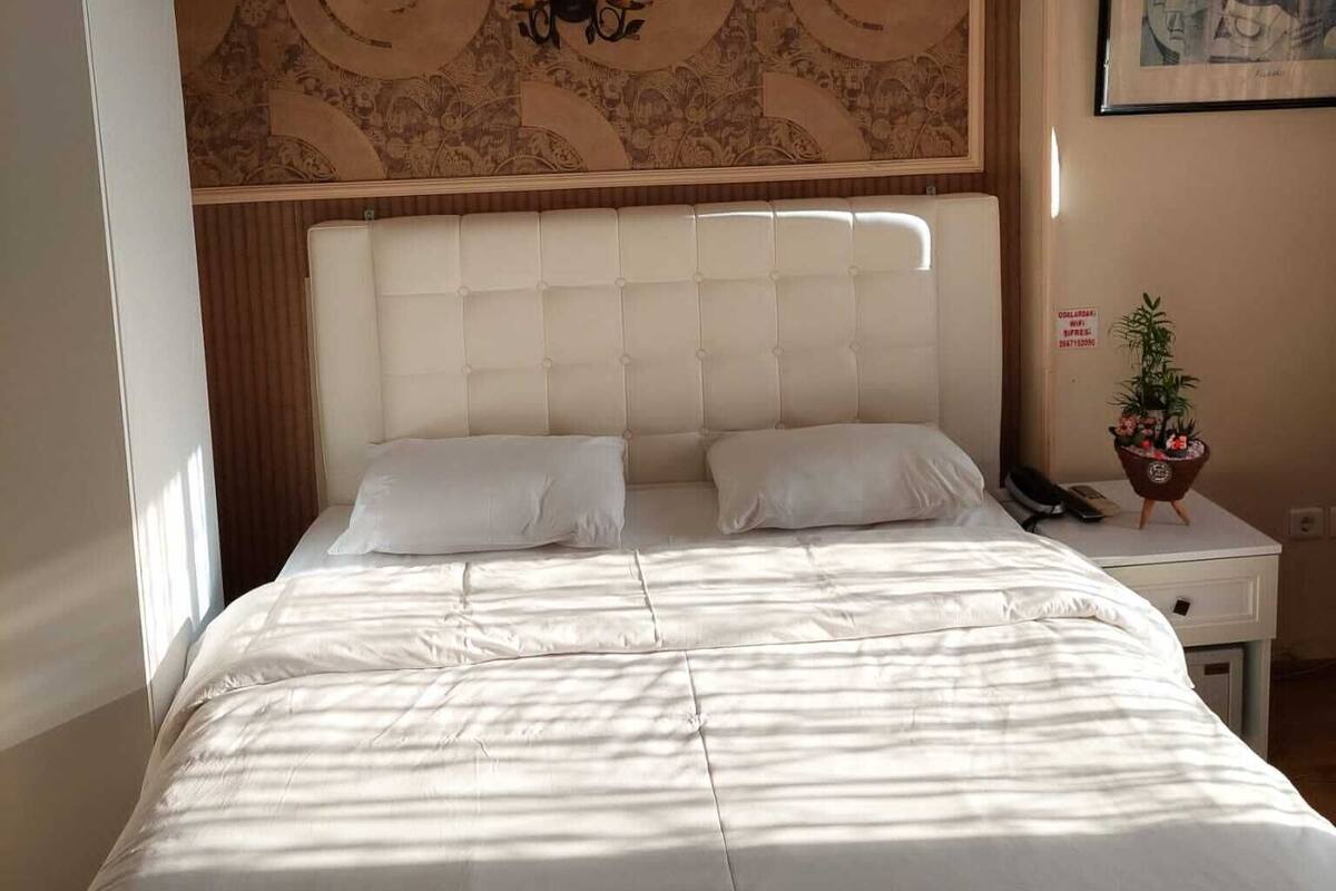 Habitación de hotel con una cama grande, cabecera con panel decorativo, mesita de noche con teléfono y planta, y un cuadro en la pared.