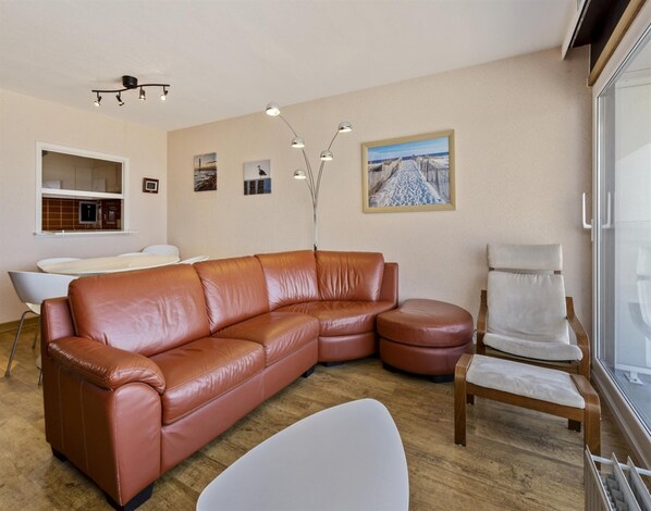 Una sala de estar moderna con un sofá de cuero marrón, un sillón blanco y una mesa de centro de madera. Hay una zona de comedor con una mesa y sillas blancas, y una cocina con una ventana.