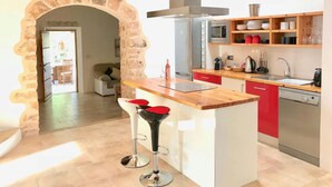 A modern kitchen with a central island, red cabinets, and a stone archway leading to a dining area. - Villa With Private Pool (Sant Cebria de Vallalta)