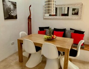 A dining area with a wooden table, white chairs, and a fruit bowl. There is a sofa with red and black cushions in the background. - Villa With Private Pool (Sant Cebria de Vallalta)