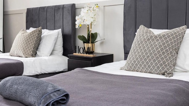 Ein Hotelzimmer mit zwei Betten, jeweils mit grauer Decke und Kissen, einem Nachttisch mit Blumenvase und einem dunklen Kopfteil.