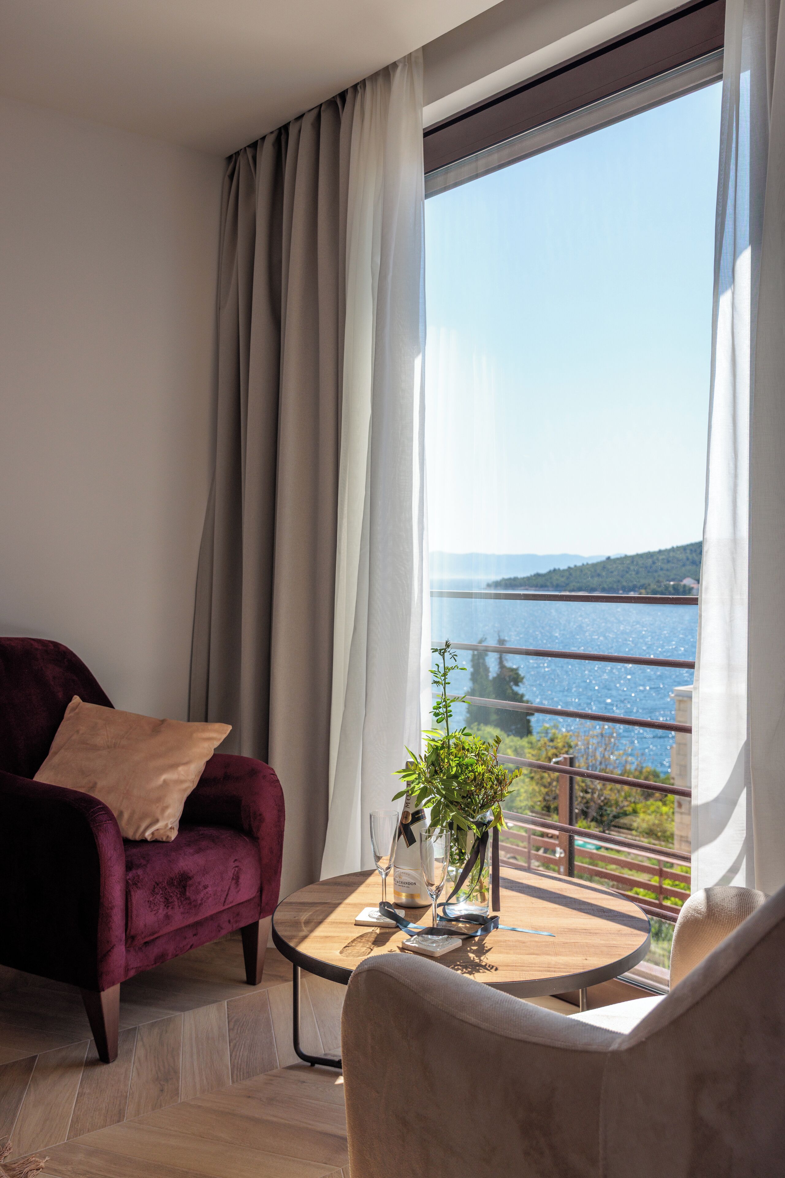 A modern living room with a view of a lake, a red velvet armchair, a round wooden table with a vase and wine glass, and a balcony railing.