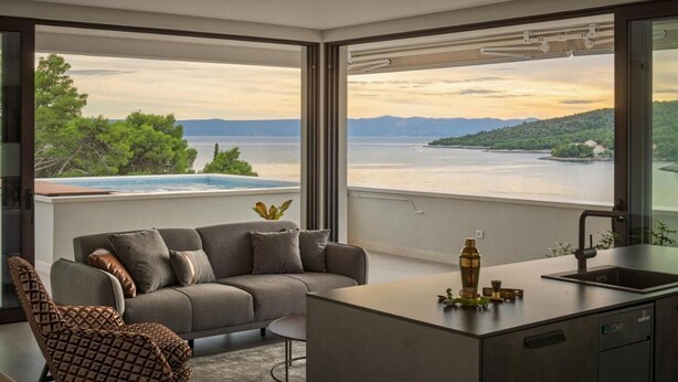 Ein modernes Wohnzimmer mit einer Couch, einem gemusterten Sessel und einem kleinen Tisch. Im Hintergrund ist ein Küchenbereich mit Spüle und einer dekorativen Flasche zu sehen. Große Fenster bieten Blick auf einen See und ferne Hügel.