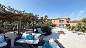 Een terras met rieten meubilair en een pergola, een zwembad met ligbedden, en een gebouw met gebogen ramen en een betegeld dak.