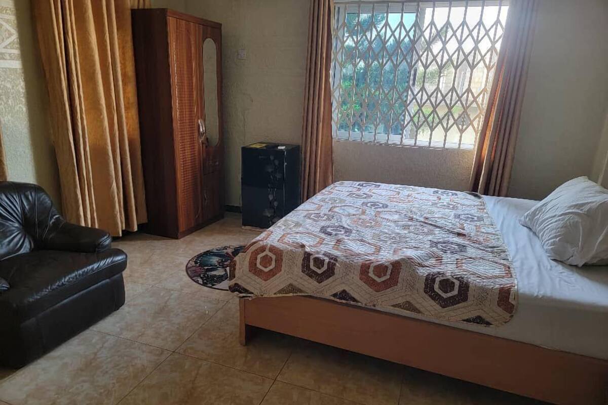 A bedroom with a bed, a black leather sofa, a wardrobe, and a window with a view of greenery.