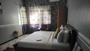 Ein Schlafzimmer mit Bett, Nachttisch, Sessel, Fenster mit Vorhängen und einer Wand mit Musterdesign.
