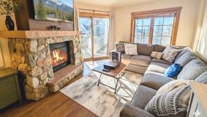 A cozy living room with a stone fireplace, a large flat-screen TV mounted above it, a comfortable sectional sofa, and a view of mountains through the windows. - Pool/Hot Tub/Views, Walk to River Run Festivals (Keystone)