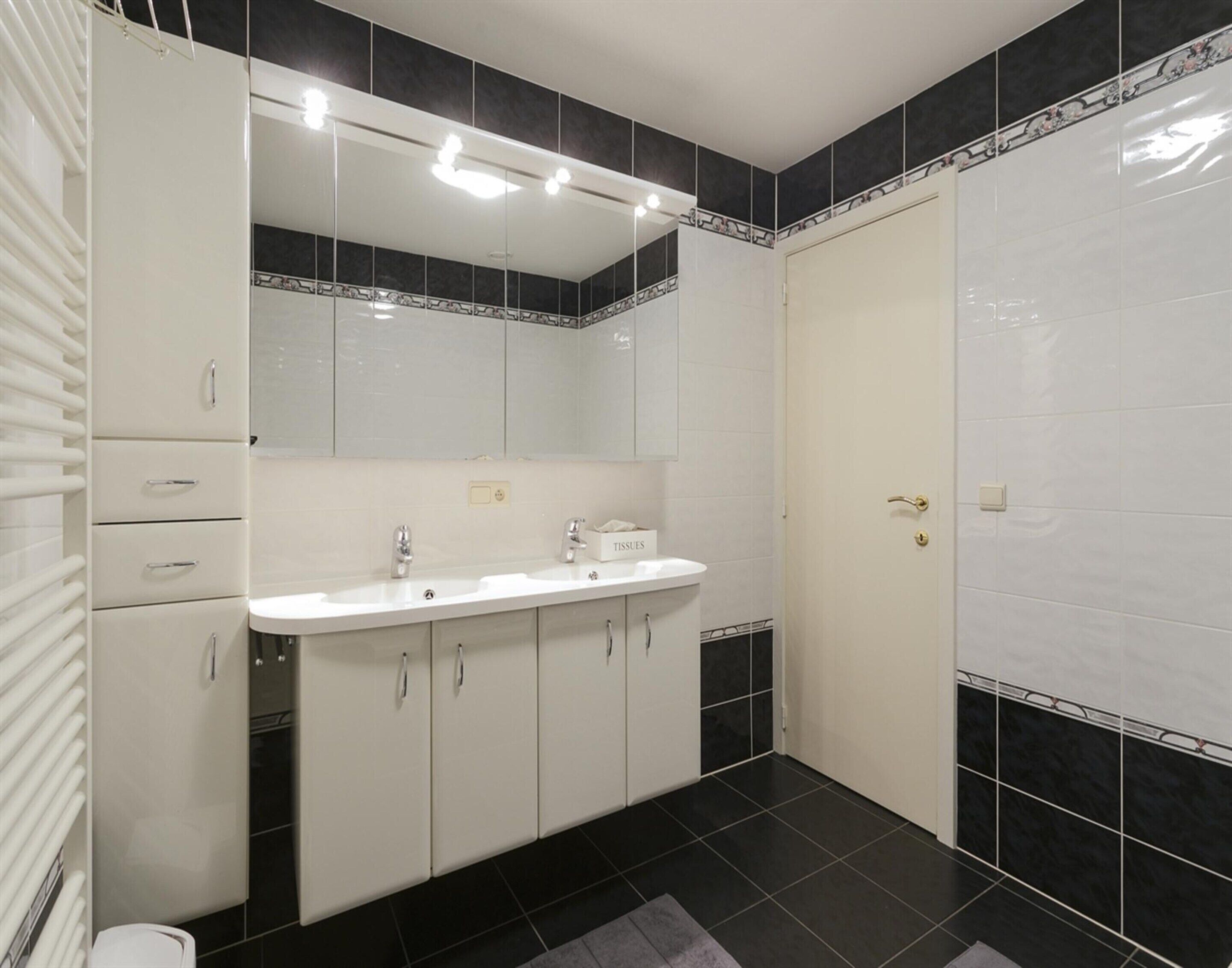 Un baño con lavamanos, espejo y armarios blancos, paredes de azulejos negros y una puerta.
