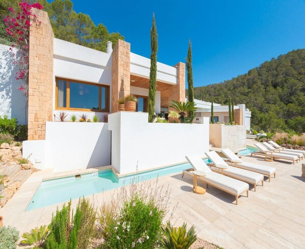 A modern villa with a swimming pool, sun loungers, and a mountainous backdrop.