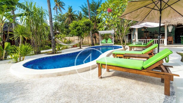 A pool area with lounge chairs, a thatched-roof structure, and tropical vegetation. - Ings Garde (Gili Air)