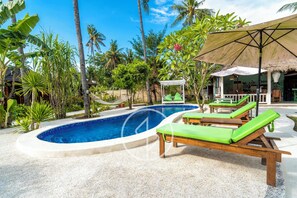 A pool area with lounge chairs, a thatched-roof structure, and tropical vegetation. - Ings Garde (Gili Air)