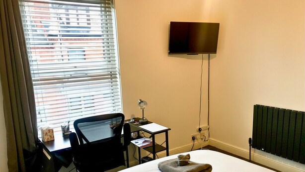 A hotel room with a bed, a desk with a chair, a television mounted on the wall, and a window with blinds.