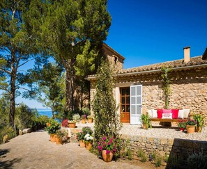 A stone house with a tiled roof, a wooden door, and a bench with cushions outside.