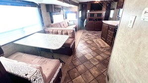 Interior compacto de um veículo recreativo, com cozinha compacta, espaço de refeições e área de estar com mesa e bancos acolchoados.