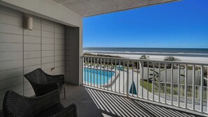 A balcony with a view of the ocean, a pool, and outdoor seating.