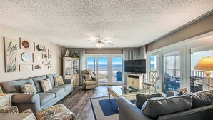 A spacious living room with a grey sofa, a coffee table, and a TV. There is a balcony with a view of the ocean.