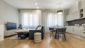 A modern living room with a sofa, dining table, and chairs, a TV, and a kitchen area with cabinets and a countertop. - HouSmart Massarenti 61 (Bologna)