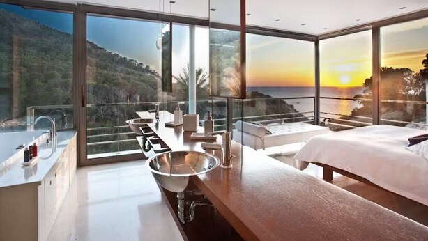 Camera d'albergo moderna con un'ampia balconata in vetro, un lavandino spazioso, un letto e vista sul mare al tramonto.