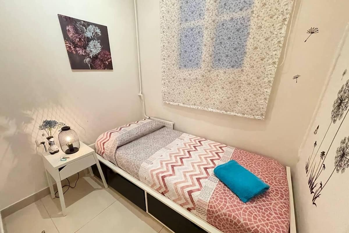 A small, neatly arranged bedroom with a single bed, a bedside table with a lamp, and a wall-mounted artwork.