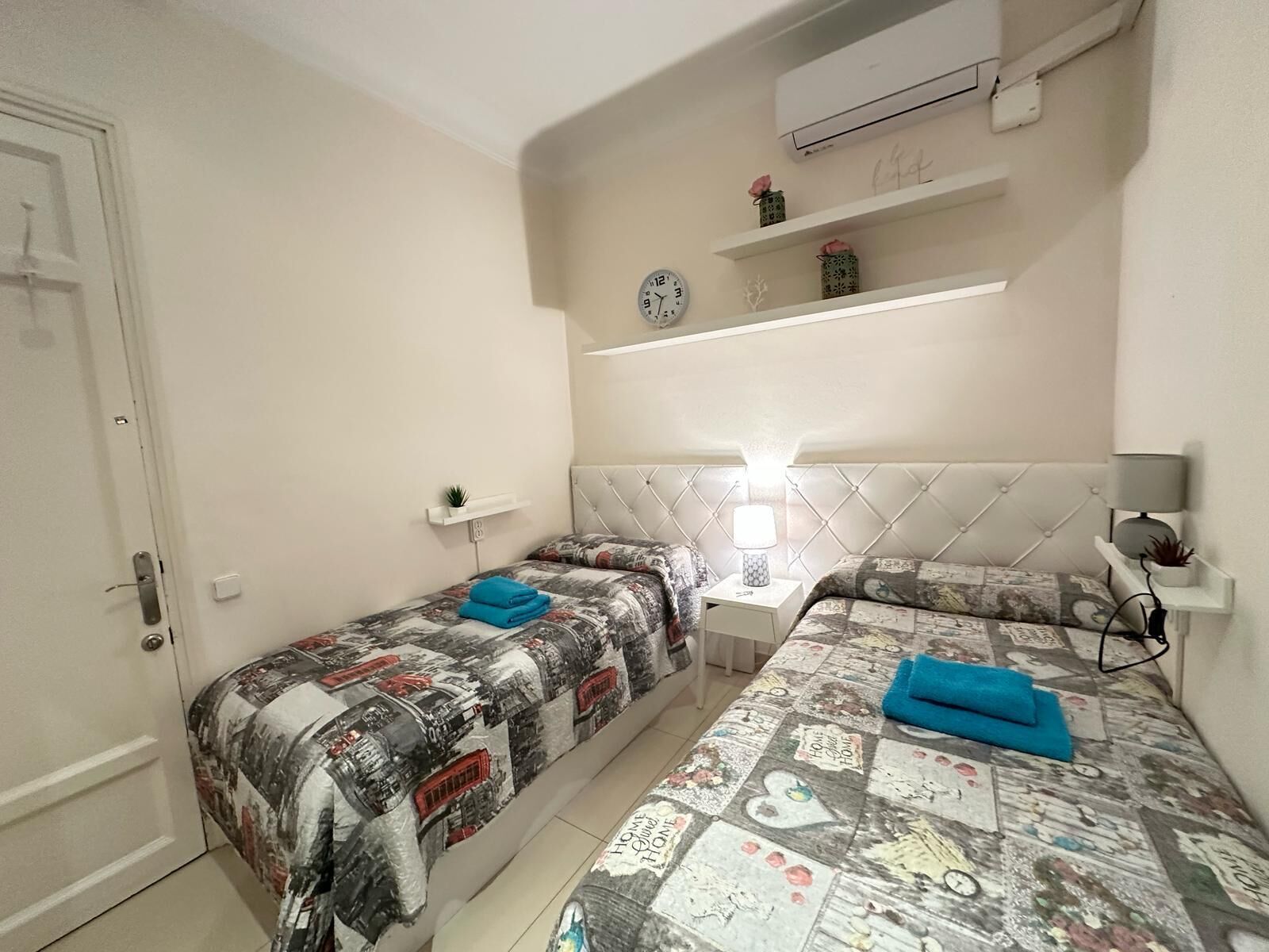 Una habitación pequeña y limpia con dos camas, una mesita de noche con lámpara, aire acondicionado en la pared y un estante con objetos decorativos.