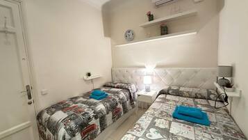 Una habitación pequeña y limpia con dos camas, una mesita de noche con lámpara, aire acondicionado en la pared y un estante con objetos decorativos.