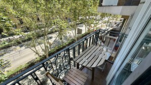 Een balkon met een houten bankje en metalen hekwerk, met uitzicht op een straat met bomen en gebouwen.