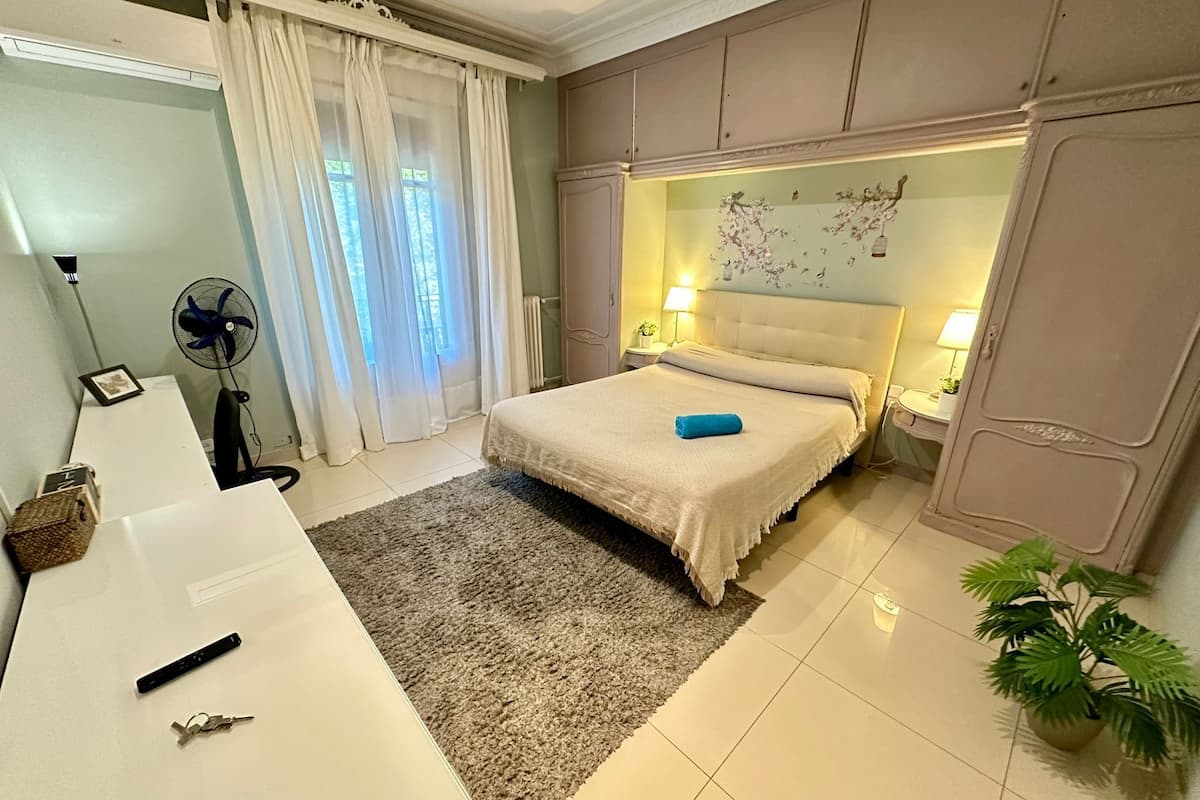A bedroom with a bed, bedside tables, a desk, a fan, a plant, and a window with curtains.