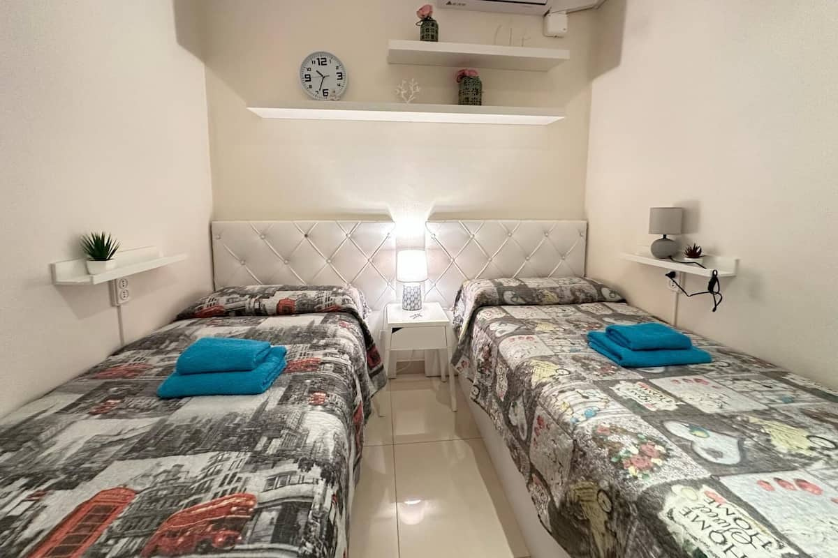 A small, well-lit room with two beds, each with a patterned bedspread and a blue pillow. There are white shelves with decorative items and a wall-mounted clock.