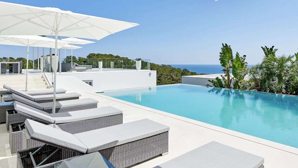 Una piscina infinita en la azotea, con sillones de descanso blancos y sombrillas, con vistas a una playa y el océano.