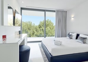 A modern hotel room with a large bed, a desk, and a view of trees through large windows. - Stylish Villa Near Ibiza Town (Ibiza Town)