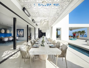 A modern dining area with a long table set for a meal, surrounded by wicker chairs, under a decorative ceiling with intricate patterns. - Exclusive Villa on White Isle (Llucmajor)