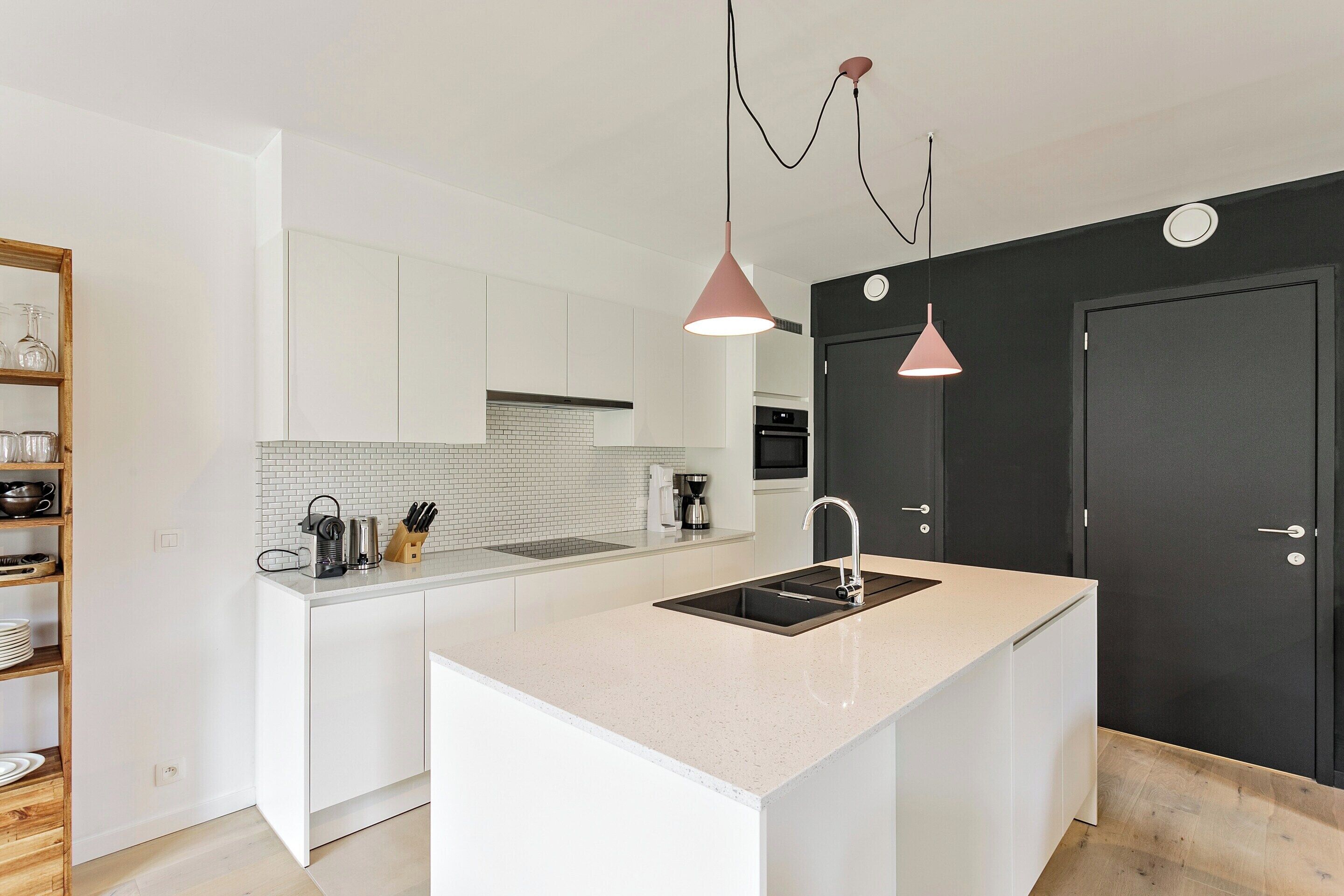 Une cuisine moderne avec des meubles blancs, un îlot central doté d’un évier et des luminaires suspendus.