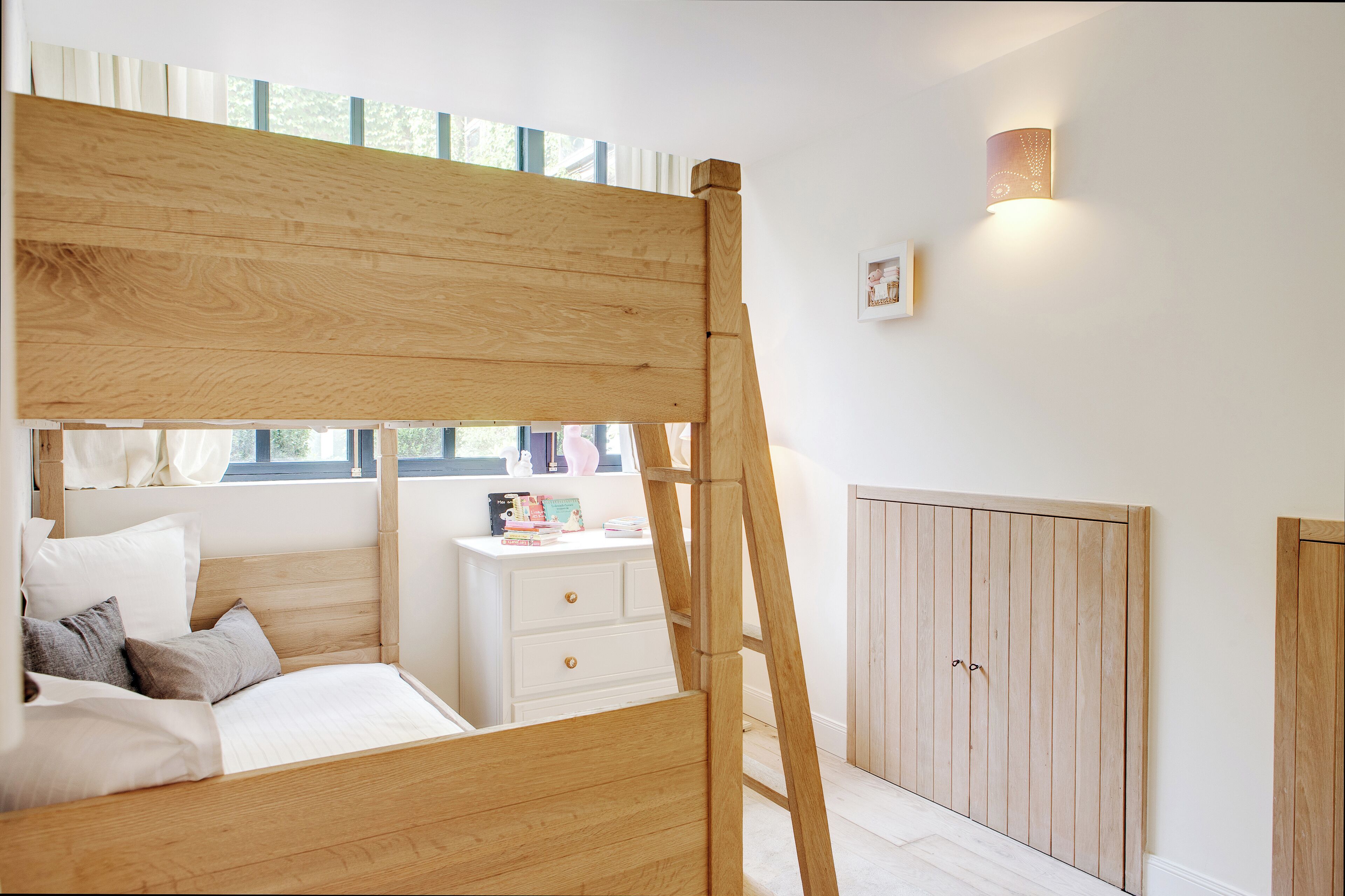 A bedroom with a wooden bunk bed, a bedside table with books, a wardrobe, and a window with curtains.