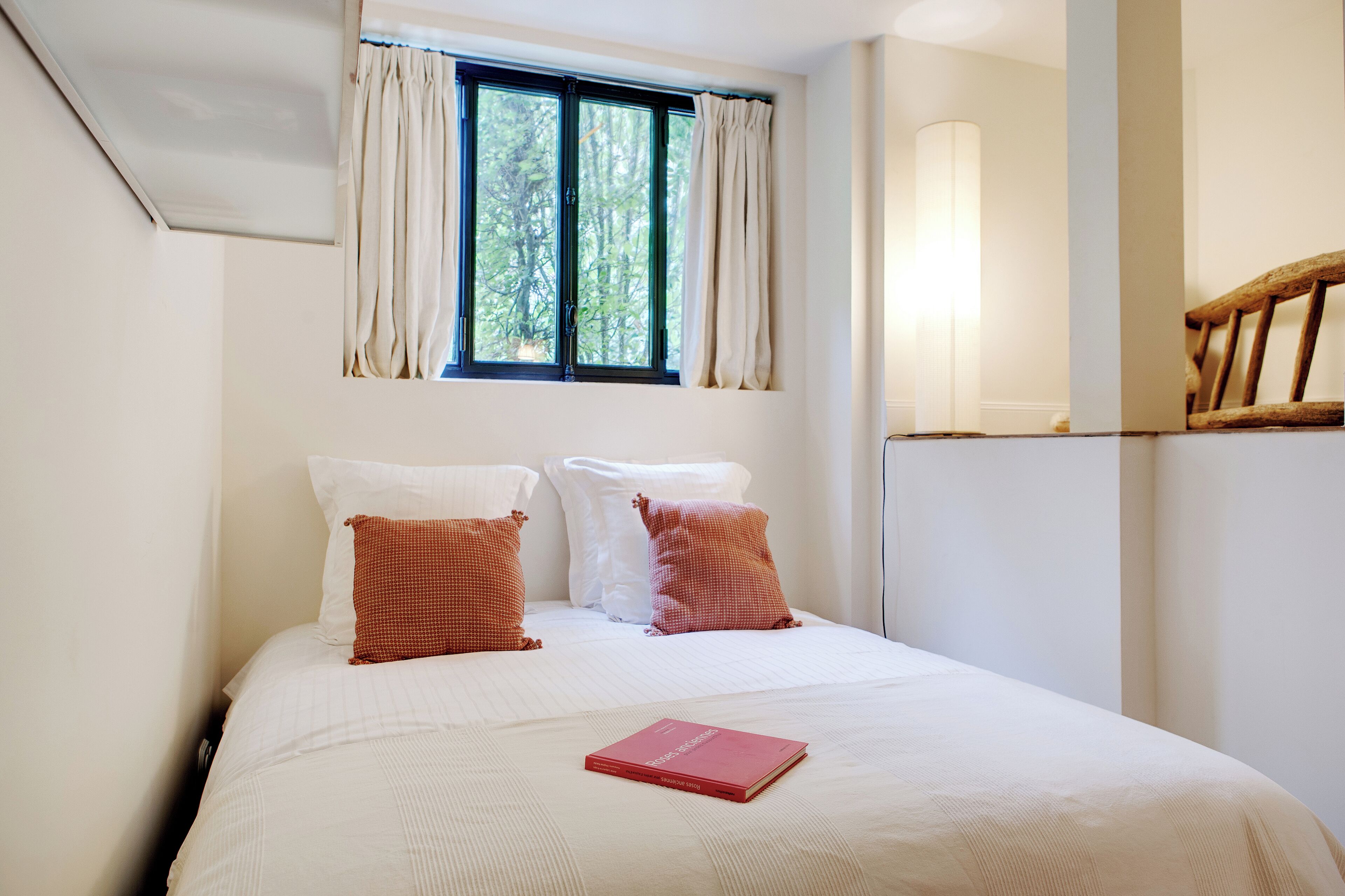 A neatly made bed with white and red pillows, a red book on the bed, and a window with curtains.