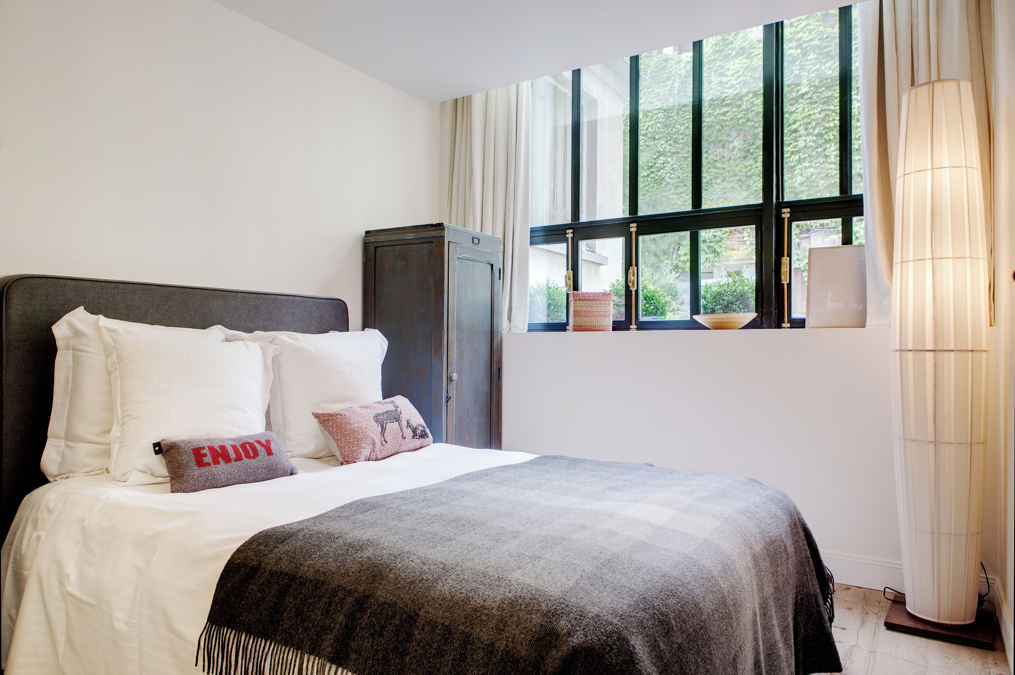 Een netjes opgemaakt bed met wit en grijs beddengoed, een donker hoofdbord en decoratieve kussens, waaronder één met de tekst 