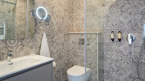 Un baño con lavamanos, espejo y retrete. Las paredes tienen azulejos con diseños y una cabina de ducha de vidrio.