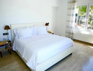 A large bed with white bedding, a wooden nightstand with a lamp, and a window with curtains. - Villa With Golf-course Views (Santa Eulalia del Rio)