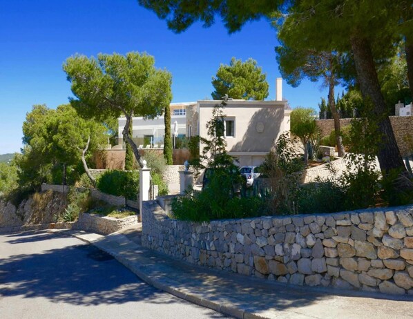 A two-story house with a stone wall and trees in the foreground. - Villa With Golf-course Views (Santa Eulalia del Rio)