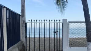 Ein Strandtor mit einem Metallzaun, Palmen und freiem Blick auf das Meer.