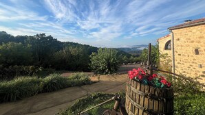 A stone building with a rustic appearance, a pathway leading to a wooden barrel with flowers, and a view of a lush green landscape under a clear sky. - La chambre du poete (vernon)