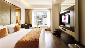 A hotel room with a large bed, a TV, a window with a city view, and a small table with a lamp.