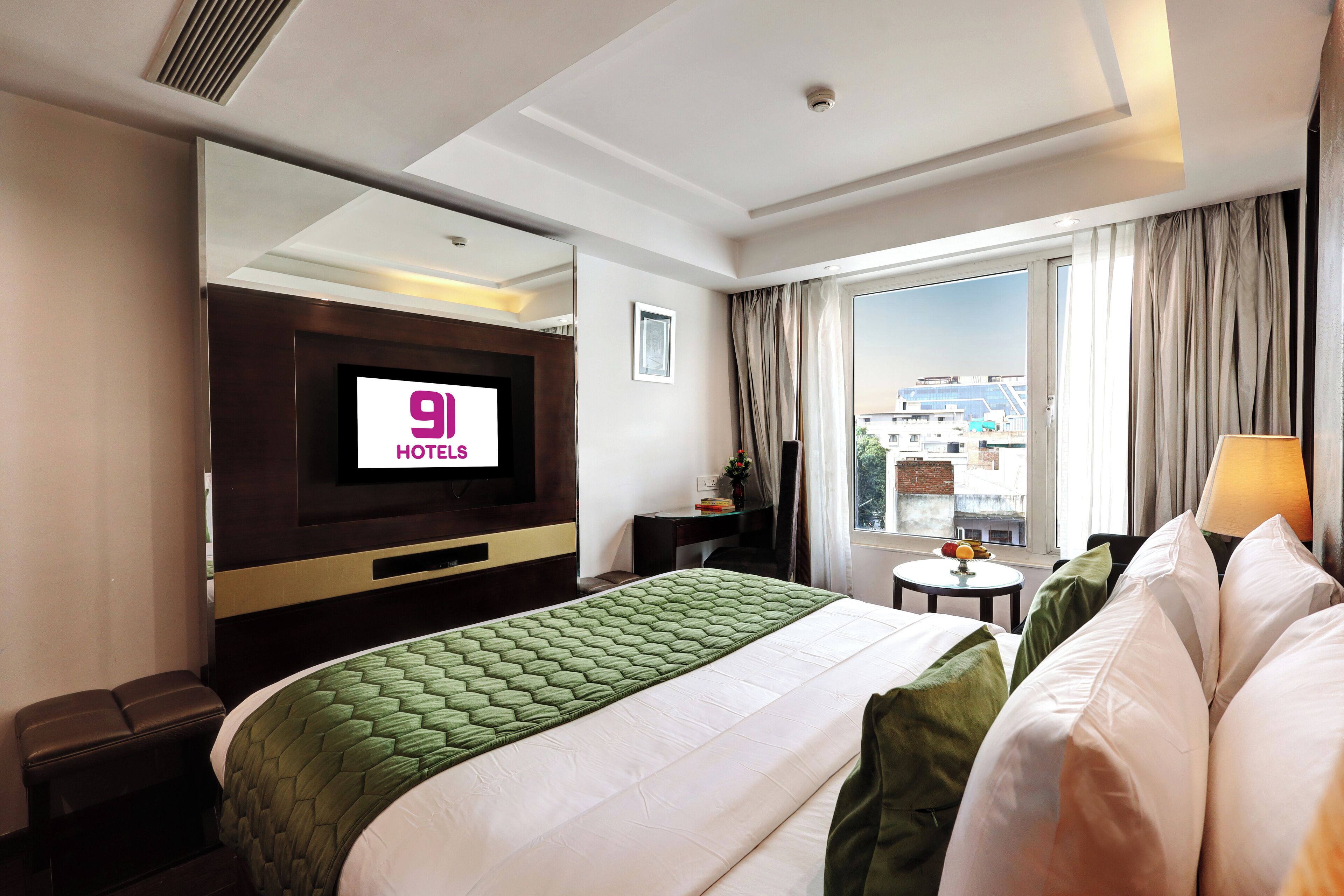 Habitación de hotel con cama, televisión, escritorio, silla, ventana con cortinas y vistas a edificios.