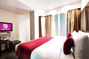Quarto de hotel com cama, televisão, escrivaninha com chaleira e janela com cortinas.