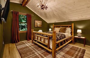 A wooden-framed bed with a patterned bedspread, a window with red curtains, a chandelier, and a small table with a lamp. - Easy Access •Mtn View• Hot Tub• Pool• Game Room• Porch• Wildlife•Minutes to Town (Ellijay)