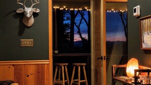 A room with a mounted deer head, wooden stools, a table with a lamp, and a painting on the wall. - Easy Access •Mtn View• Hot Tub• Pool• Game Room• Porch• Wildlife•Minutes to Town (Ellijay)