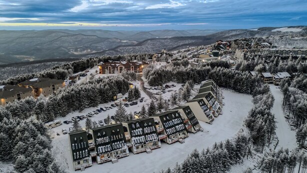 Une station de ski enneigée, composée de plusieurs bâtiments, dont un grand chalet et plusieurs petits pavillons, entourée de forêts denses et de montagnes.