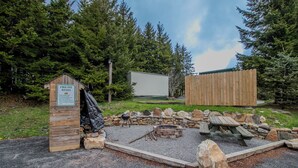 Un espace aménagé avec un foyer, une structure en bois portant l’inscription « Règles d’utilisation du foyer », un foyer en pierre et une table de pique-nique.